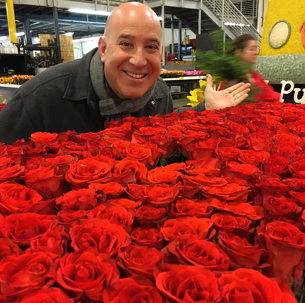 5a05e246e49a4b00015b7640_Stefan-and-Roses Man smiling behind a large display of vibrant red roses in a flower market setting.