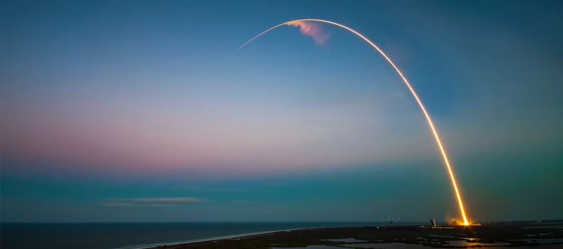 Rocket launch at dusk with curved trajectory over coastline