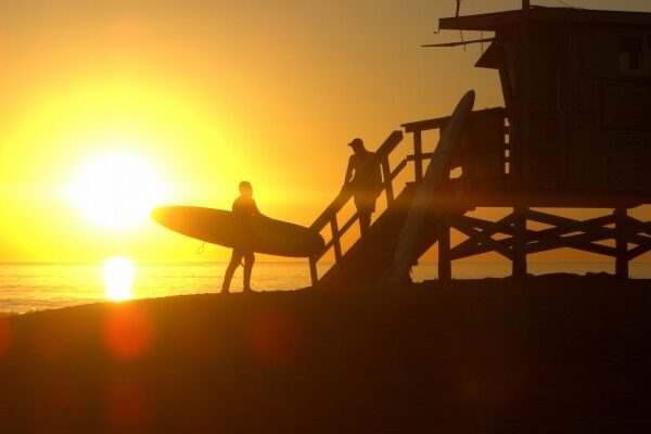 Silhouetted surfers by the beach at sunset near lifeguard tower.