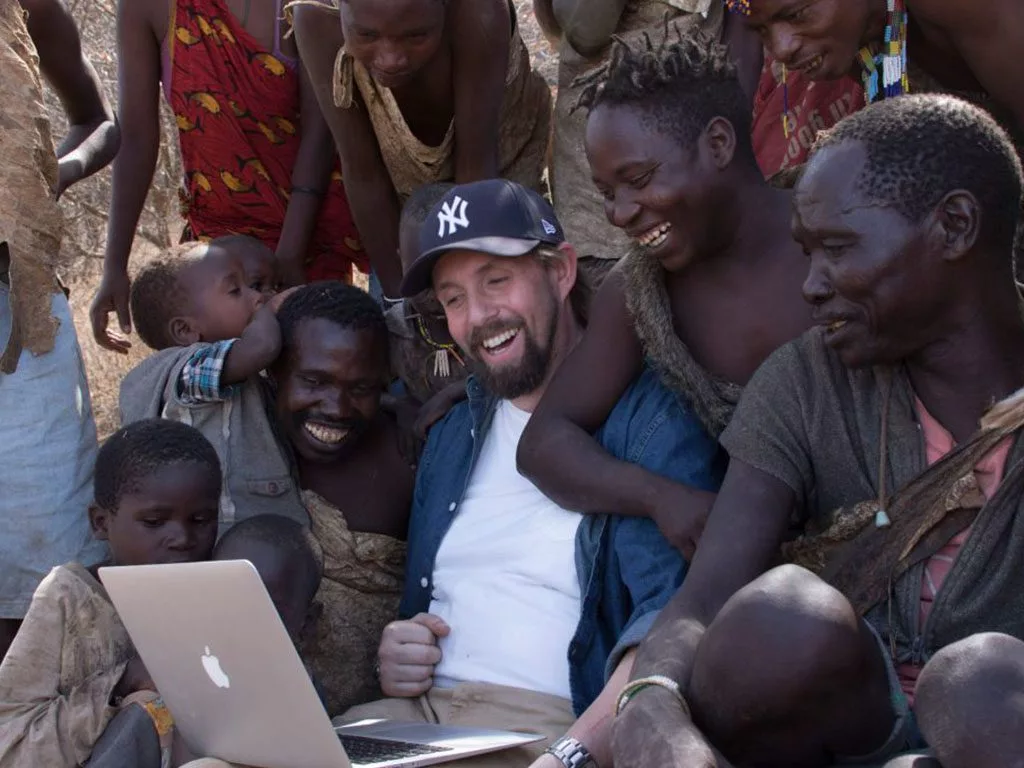 Group of diverse people laughing around a laptop in a rural setting.
