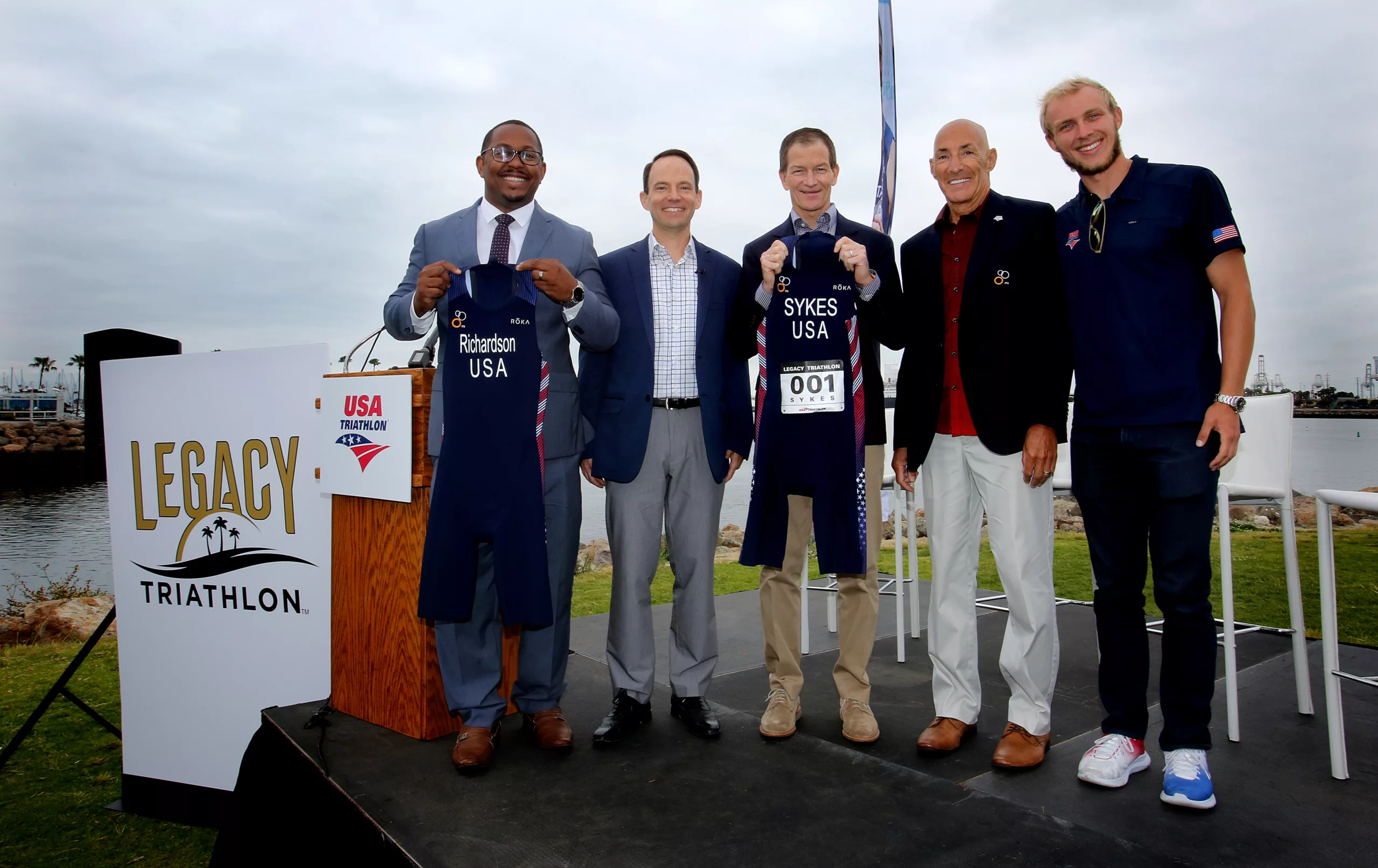 Five men at Legacy Triathlon event holding race number bibs by the sea