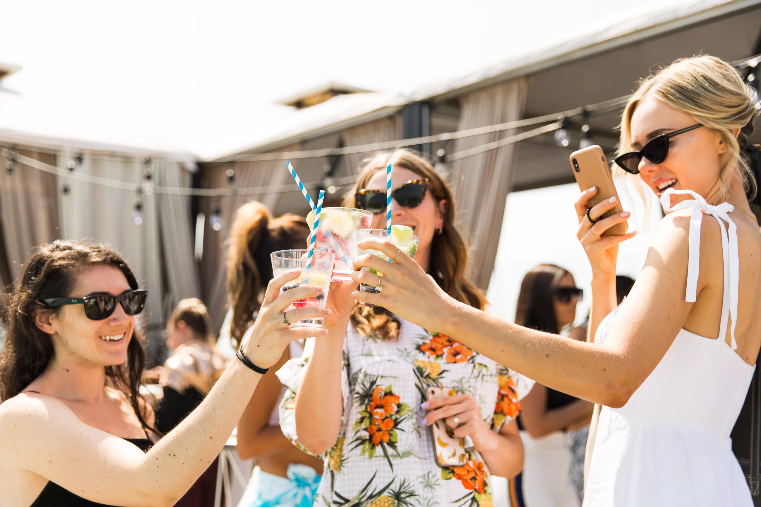 Women toasting drinks at sunny rooftop party with one using smartphone