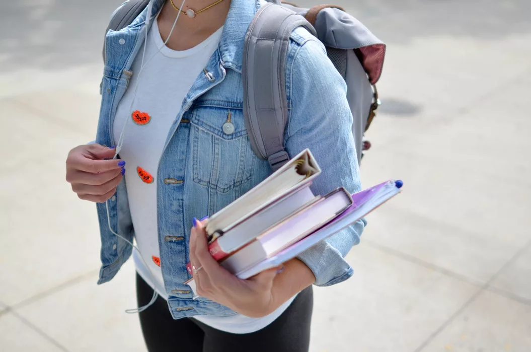 Student with denim jacket carrying textbooks and backpack outdoors