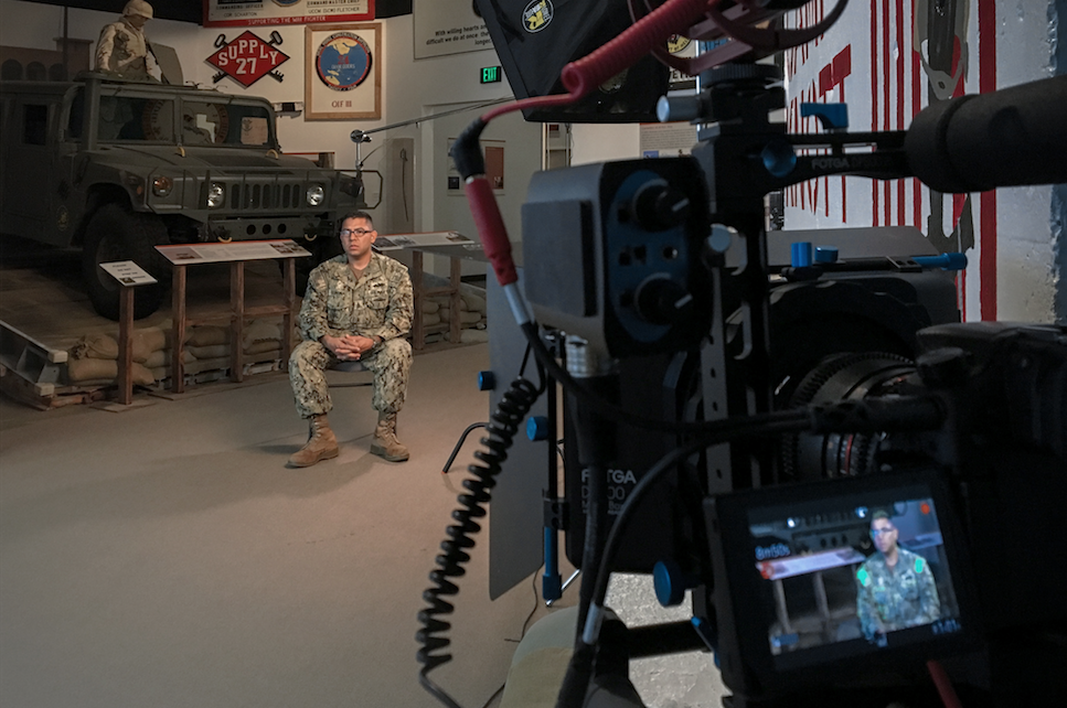 Soldier sitting for interview in museum with camera in foreground and Humvee in background