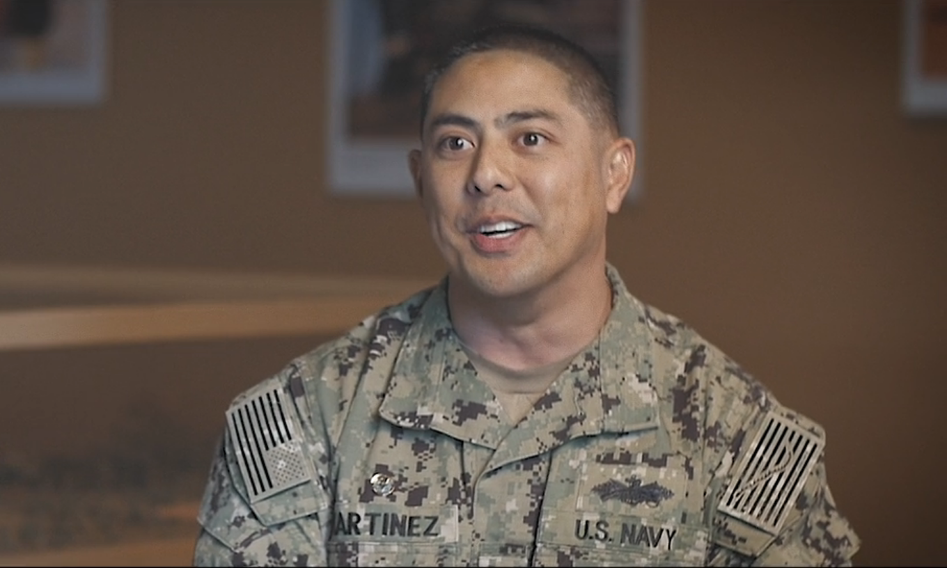 Smiling service member in U.S. Navy camouflage uniform indoors.