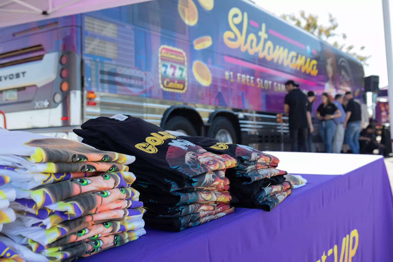 Colorful Slotomania promotional bus and stacked t-shirts at an event.