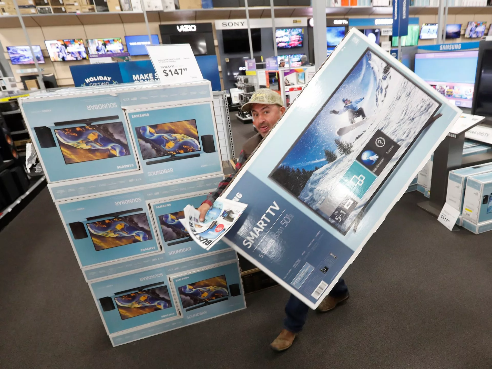 Man carrying large TV box in electronics store with TVs and soundbars on display