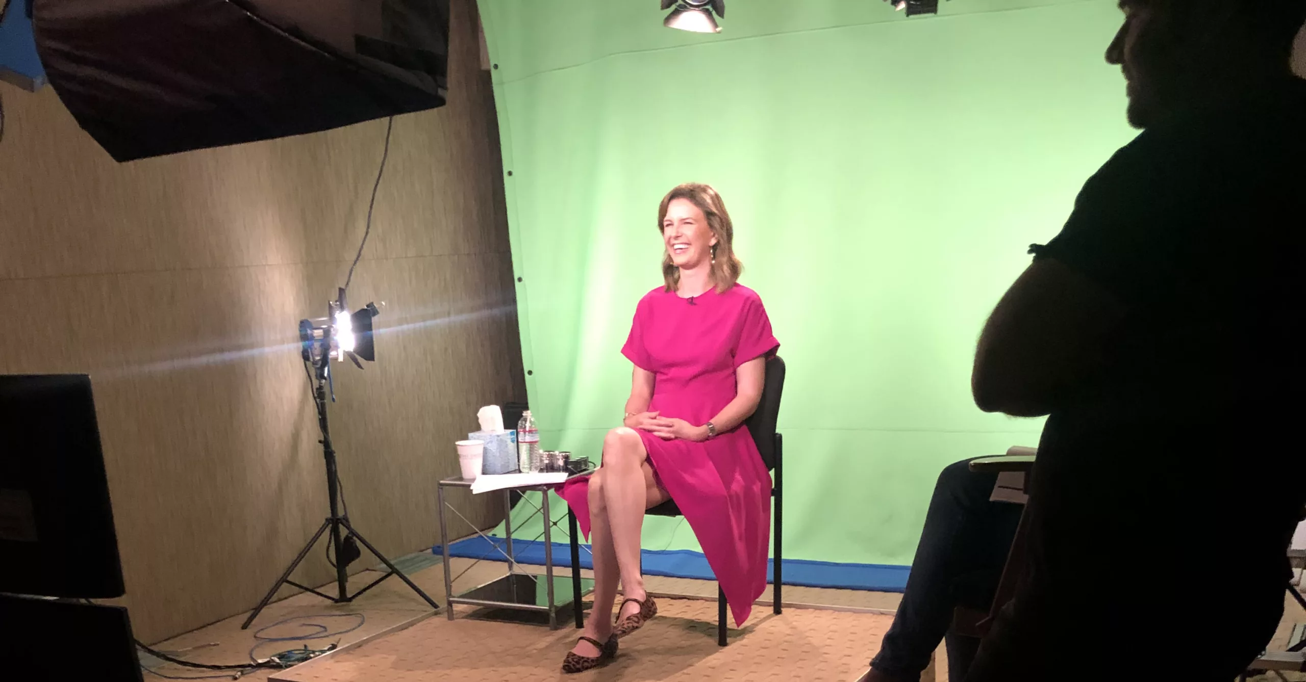 Woman in pink dress sitting for video shoot with green screen background and studio lights.