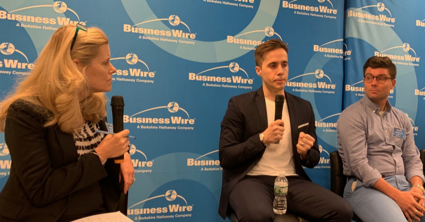 Three professionals speaking at Business Wire event with microphone and logo backdrop