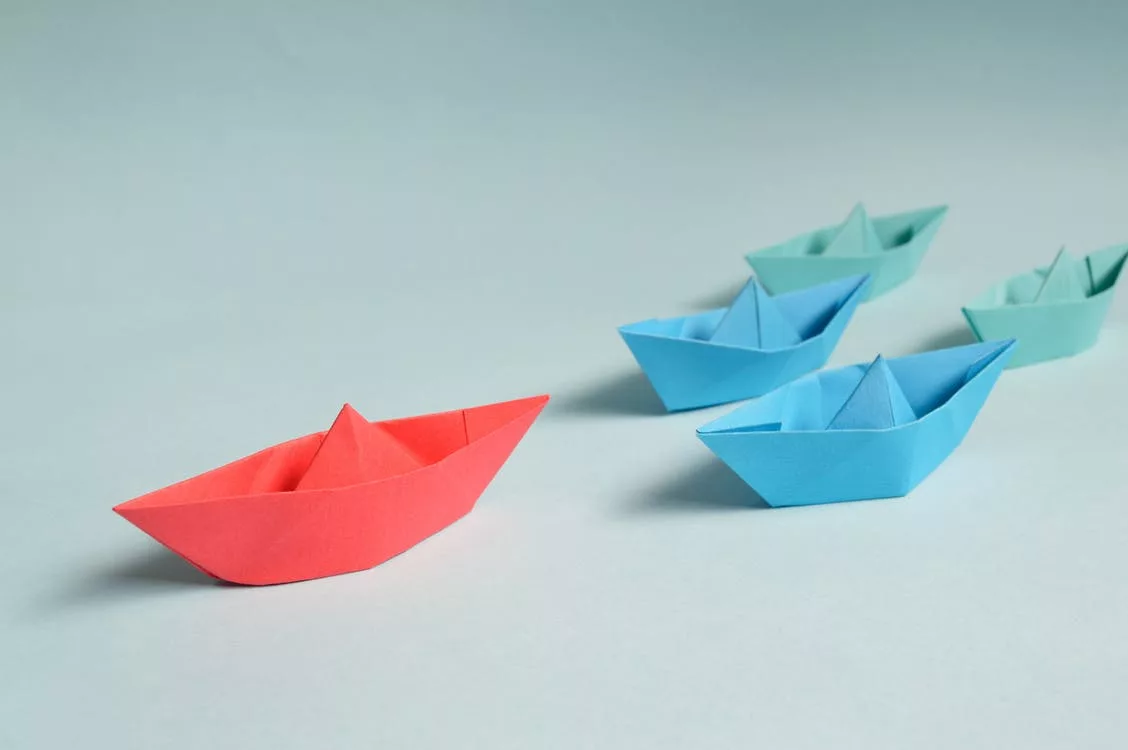 Red origami boat leading blue paper boats on a pale background