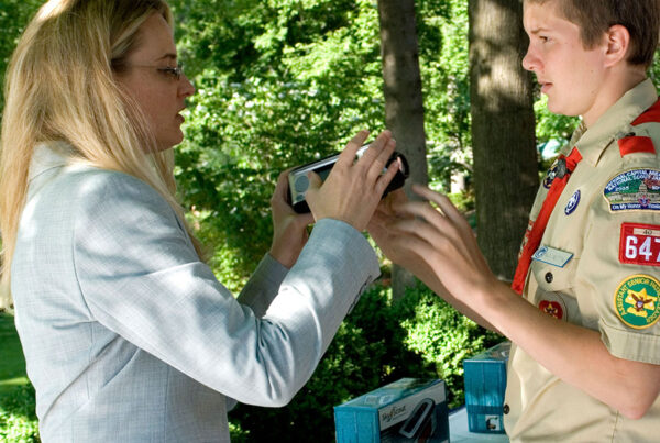 Woman in suit showing camera to boy scout outdoors near camera boxes.