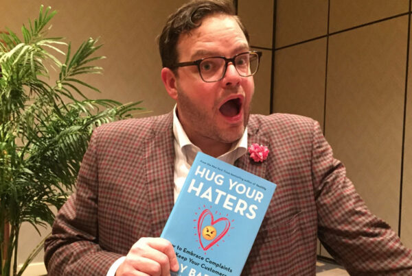 Man in suit excitedly holds "Hug Your Haters" book near potted plant.
