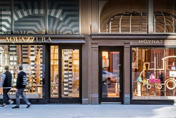People walking past Aquazzura and Moynat luxury store facades in city.