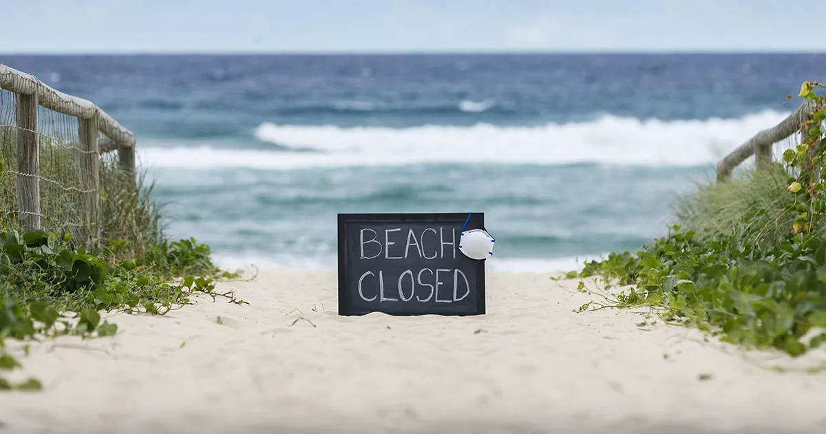 Beach closed sign with mask on sandy path by the ocean