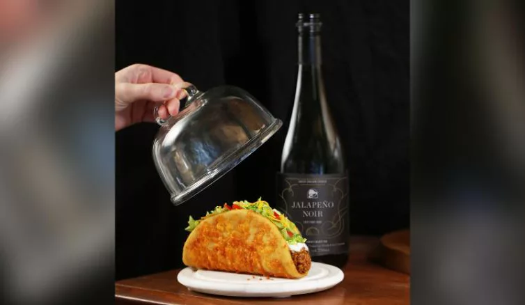 Hand lifting silver cloche from taco with lettuce and wine bottle in background