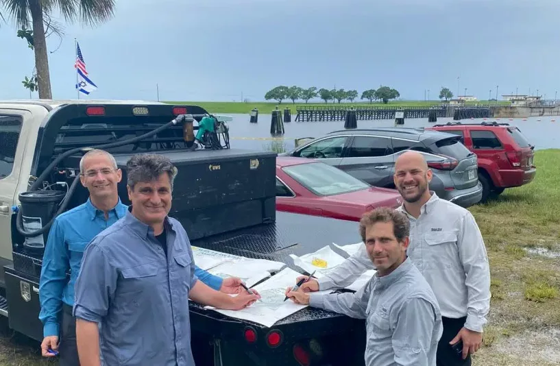 Four men reviewing plans on truck bed by waterfront with parked cars