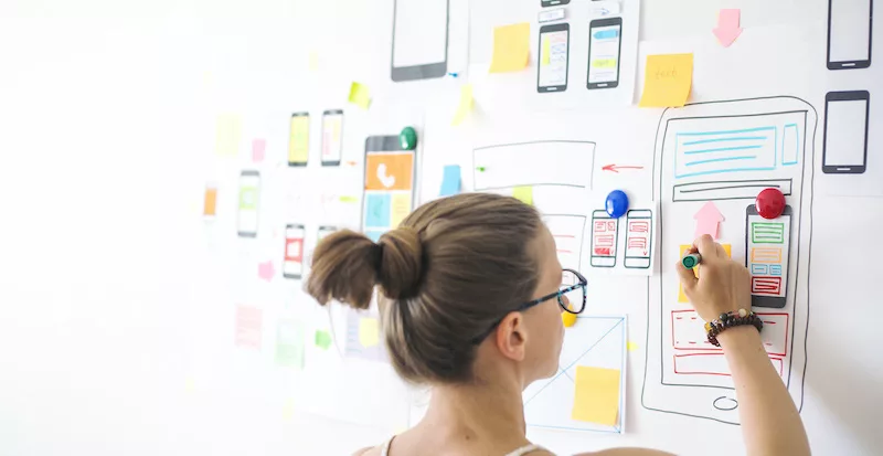 Woman with glasses drawing a mobile app UI/UX design on a whiteboard with sticky notes.