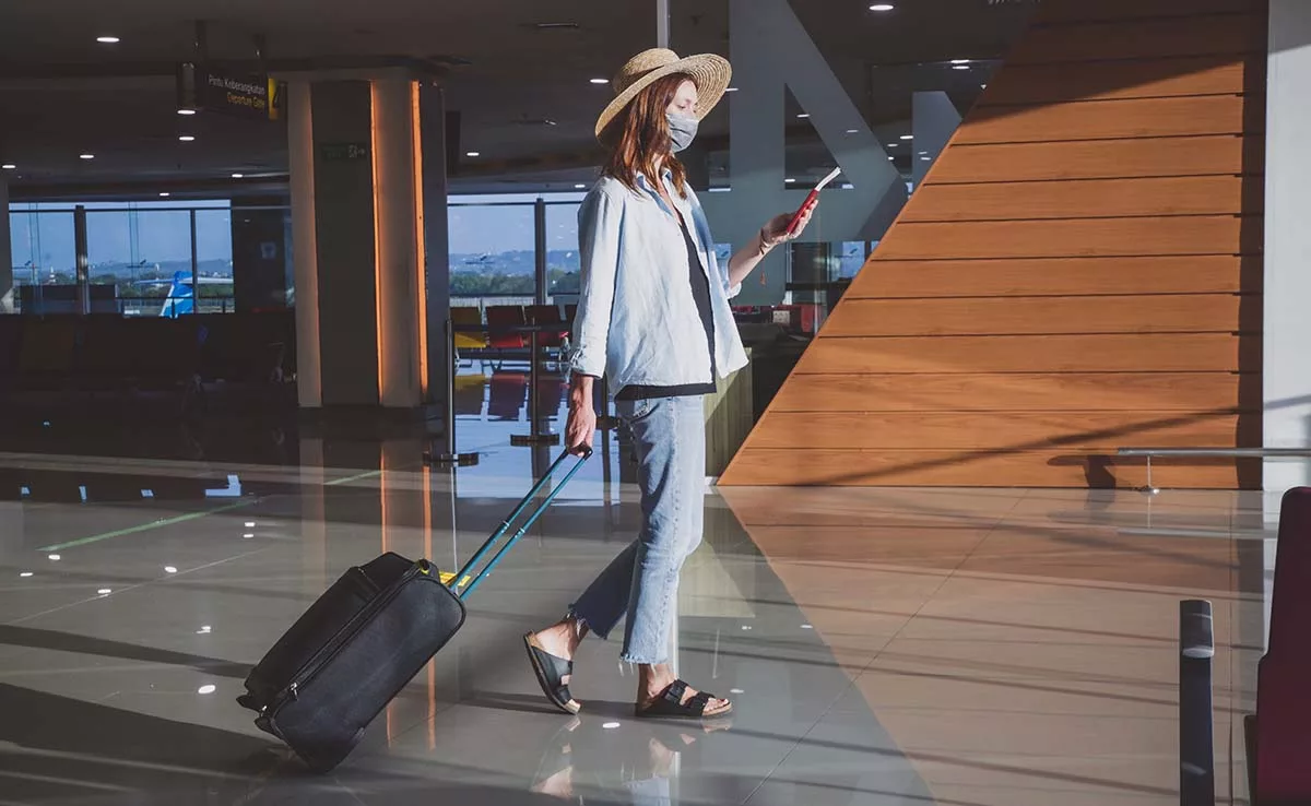 Woman with hat and mask pulling suitcase in airport lounge looking at phone