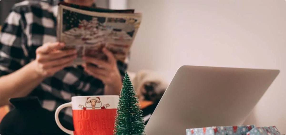 Person reading newspaper with holiday mug and mini Christmas tree on desk near laptop.
