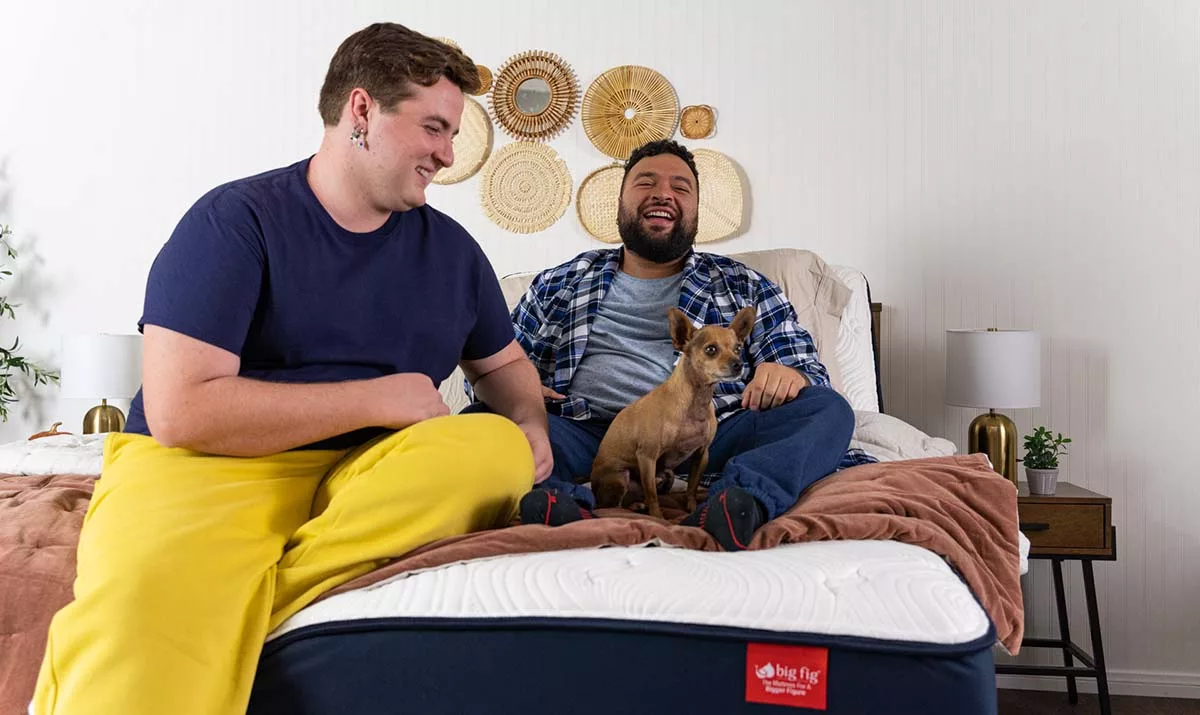 Two men and a dog sitting on a bed with decorative wall art behind them.