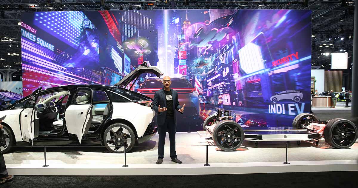 Man presenting electric car with Times Square backdrop at auto show.