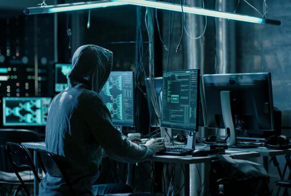 Hacker with hoodie typing on keyboard in a dark room with multiple computer screens.