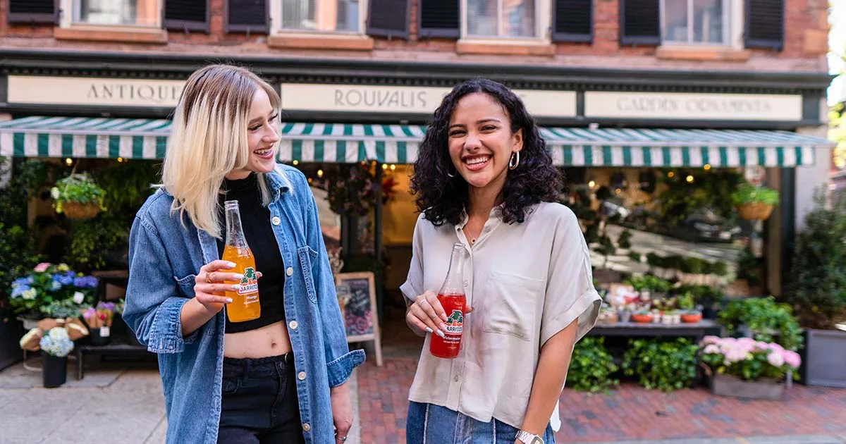 Two women laughing and holding soft drinks in urban outdoor setting