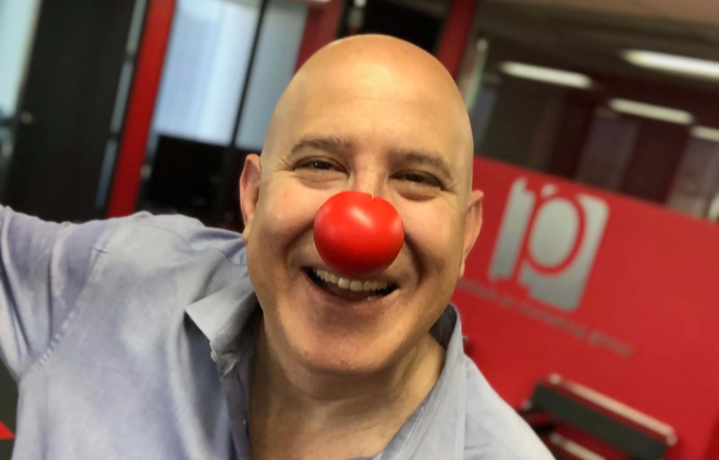 Bald man smiling with a red clown nose in an office environment
