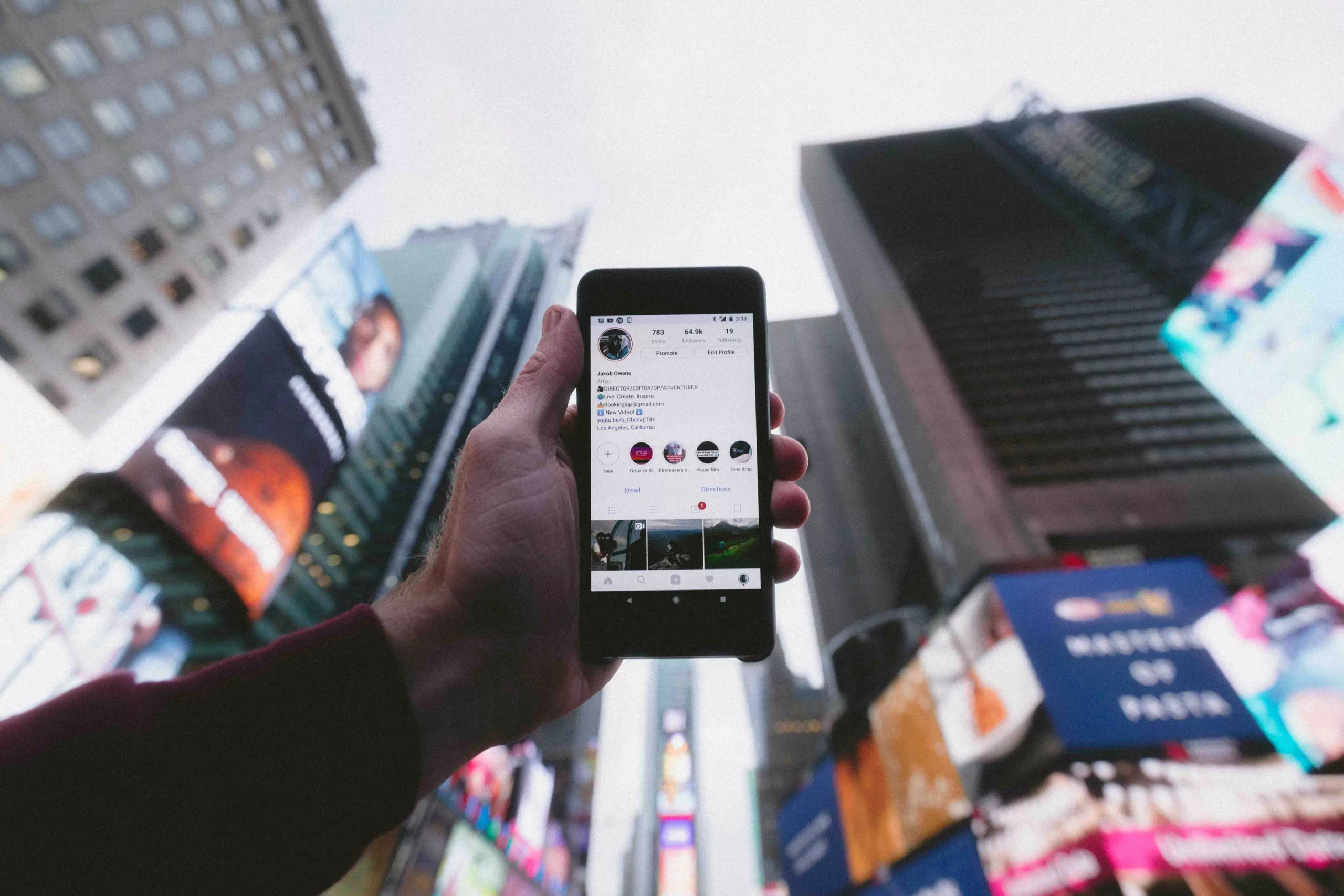 Hand holding smartphone with social media profile open against blurry cityscape with billboards.