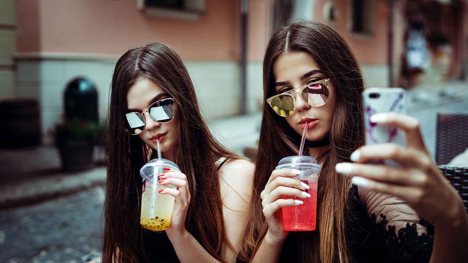 Two women in sunglasses taking a selfie while drinking colorful beverages