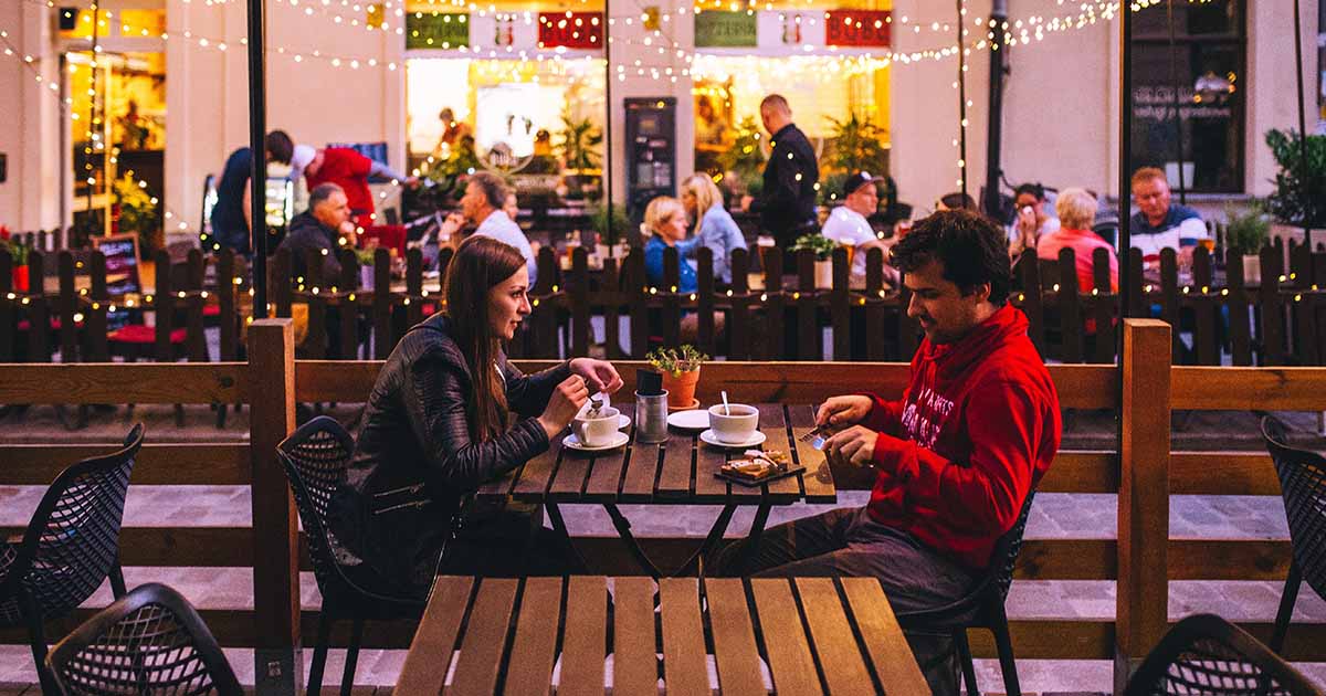 Couple enjoying coffee at an outdoor cafe with festive lights in the evening.