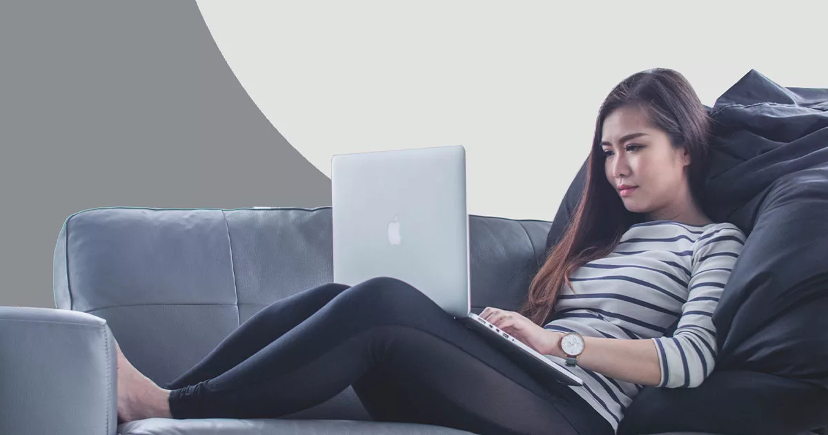 Woman working on laptop while sitting on a grey couch.