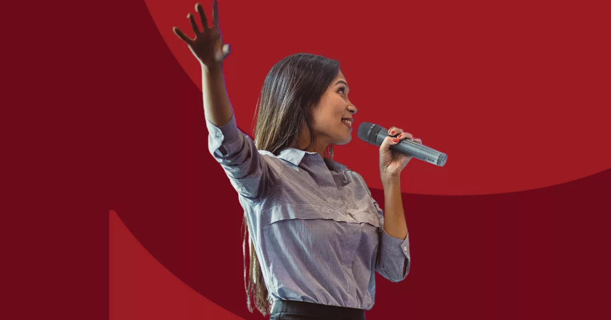 Woman singing into microphone with raised hand against red background.