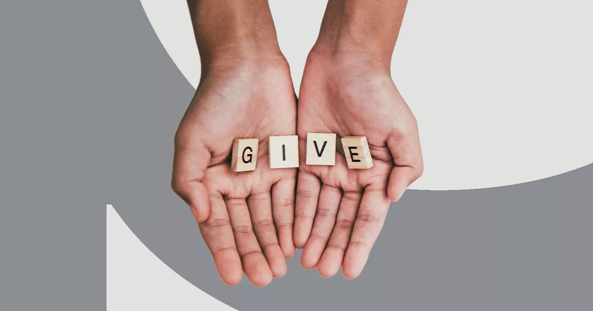Open hands holding wooden tiles spelling "GIVE" on a neutral background.