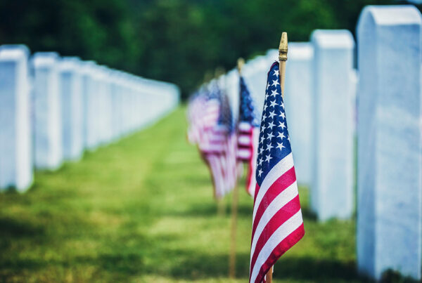 American flags lined up in a cemetery with white headstones on a sunny day
