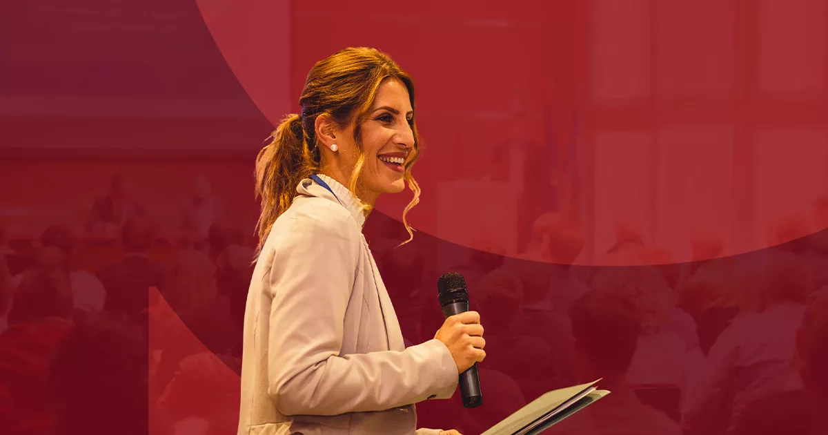 Smiling woman presenting at conference with microphone in red-themed room