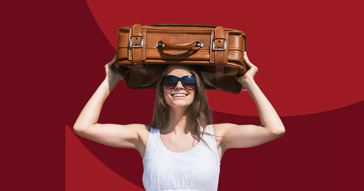 Woman smiling with sunglasses holding a brown leather suitcase overhead against a red background.