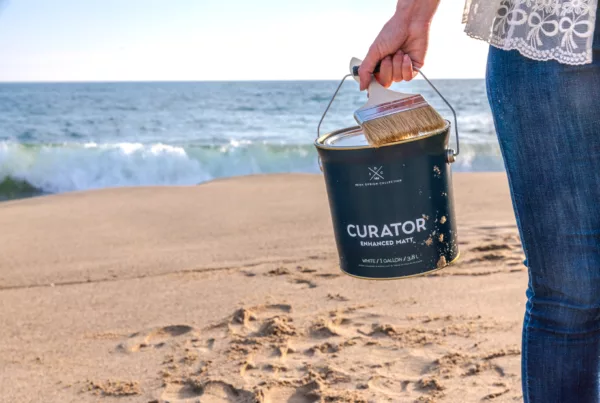 Person holding paint can and brush on beach with waves in background