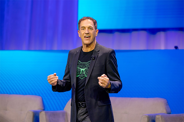 Man in a black blazer and t-shirt delivers an engaging speech on a brightly lit stage at a conference.