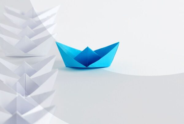 Blue paper boat moving ahead of a line of white paper boats.