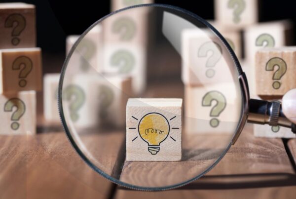 Magnifying glass hovering over a wooden block with a light bulb on it with other wooden blocks with questions marks on them behind it.