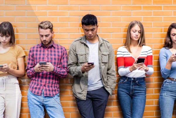 Five young adults using their cell phones while leaning against a brick wall.