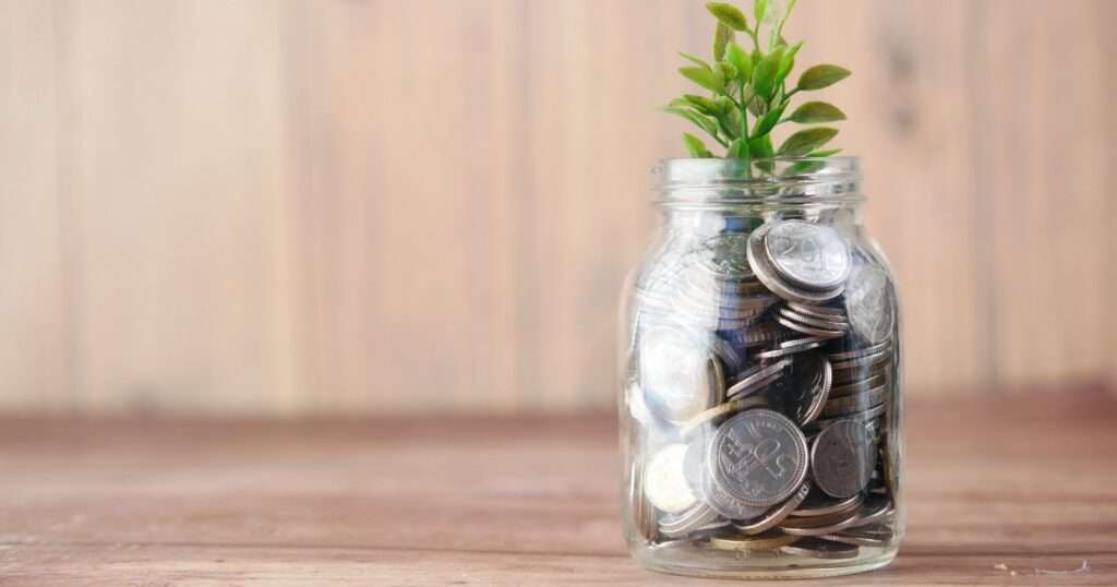 A mason jar of coins with a small plant growing out of the top against a wooden background.